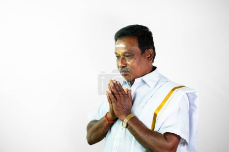 Téléchargez les photos : Un Indien du sud dans la cinquantaine portant un dothi blanc traditionnel et une chemise blanche salue avec le geste vanakkam. Il lève les mains dans une pose de namaste avec des paumes - en image libre de droit