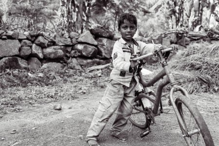 Téléchargez les photos : Un pauvre petit garçon en haillons se tient dans une rue poussiéreuse du village, tenant un vélo cassé. Il a l'air triste et abattu. L'image est en noir et blanc. - en image libre de droit