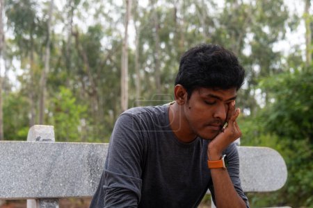 Foto de Un indio deprimido se sienta en un banco en un parque, llorando por su cara. Se ve perdido y solo, con la cabeza en las manos. - Imagen libre de derechos