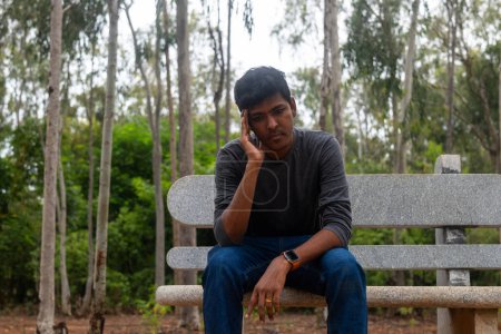 Foto de Un retrato de un indio deprimido sentado en un banco en un parque. Está mirando al suelo con una expresión triste en su rostro. Su cabello está descuidado y lleva ropa casual.. - Imagen libre de derechos