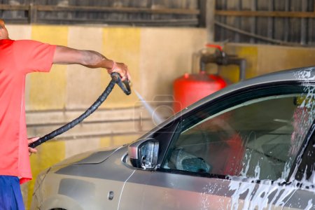 Ein Mann mit entschlossenem Gesichtsausdruck schwingt gekonnt einen Hochdruckwasserstrahl, um Schmutz aus seinem Auto in einer Selbstbedienungswaschanlage zu waschen.