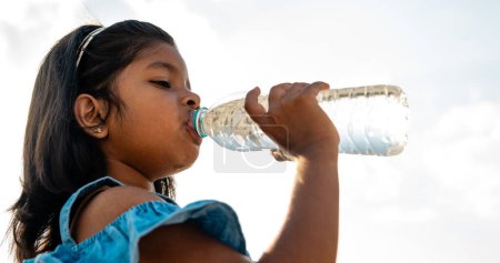 A young Asian girl enjoys a refreshing drink of water from a reusable bottle under a clear blue sky