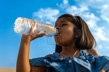 A young Asian girl takes a moment to quench her thirst with water from a reusable bottle, enjoying the bright blue sky above
