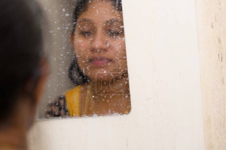 Eine nachdenkliche junge Frau steht in einem dampfenden Badezimmer und starrt ihr Spiegelbild im Spiegel an.