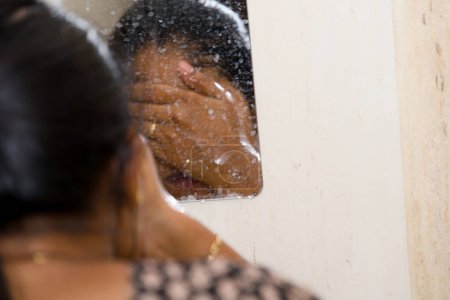 A woman gently washes her face with a cleanser in the bathroom, looking at her reflection in the mirror