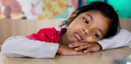 Photo for A thoughtful young Asian girl lies on her stomach with her head on a table in her room - Royalty Free Image