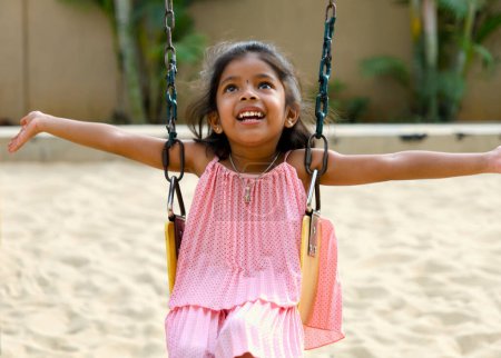 Una niña despreocupada con ojos brillantes se ríe de alegría mientras se bombea las piernas en lo alto de un columpio rojo brillante en un patio de recreo soleado