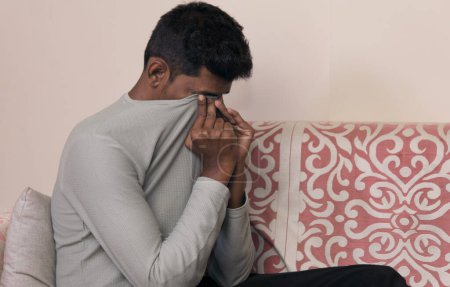 Ein Foto von einem jungen Mann, der niedergeschlagen auf einer Couch zu Hause liegt und sein Gesicht durch die Hände verdeckt.