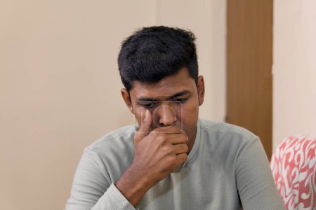 An image of a South Asian man looking downcast and wiping tears from his eyes while sitting on a couch.
