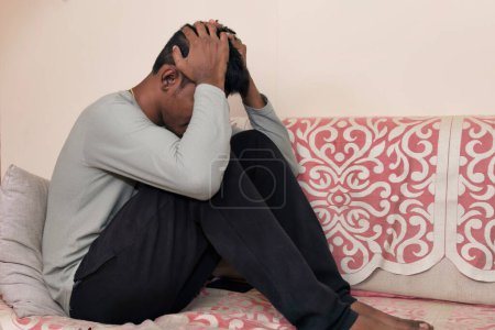 Ein indischer Mann sitzt allein auf einer Couch, den Kopf in den Händen und übermittelt Gefühle der Traurigkeit und Verzweiflung.