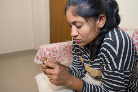 Une femme indienne réfléchie s'assoit sur un canapé dans son salon, regardant une bague à son doigt