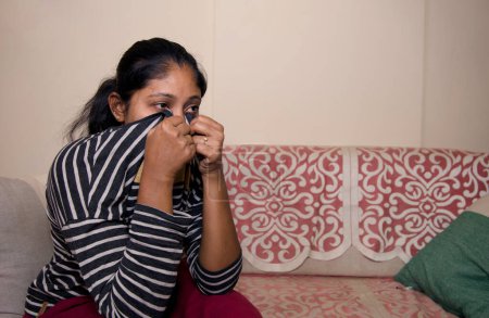 Photo for A young Indian woman sits alone at home, clutching a t-shirt to her mouth while crying - Royalty Free Image