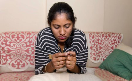A thoughtful Indian woman sits on a couch in her living room, gazing at a ring on her finger