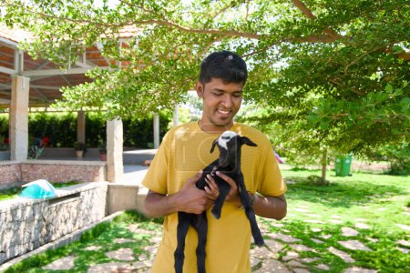 A kind-hearted Indian farmer in traditional clothing gently holds a cuddly black baby goat on a sunny farm.