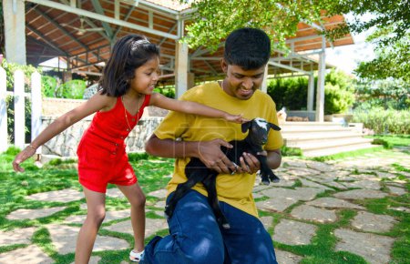 Una encantadora imagen de un padre indio y su hija creando recuerdos felices mientras juegan con una pequeña cabra encantadora en su colorido y animado jardín del patio trasero.