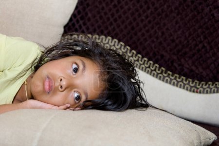 An image of a young girl lying on a couch in a living room, looking sad or disappointed.
