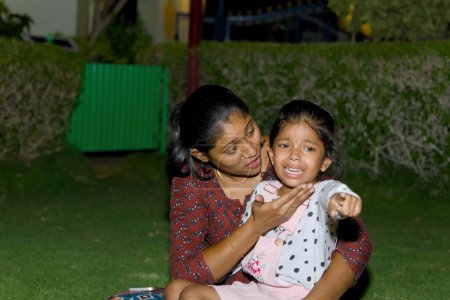Photo for A young Indian girl cries in a lush green garden while her concerned mother kneels beside her, offering a hug - Royalty Free Image