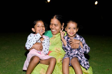 Photo for A content Indian grandmother smiles as she embraces her two granddaughters on a cozy blanket in their backyard garden at night - Royalty Free Image