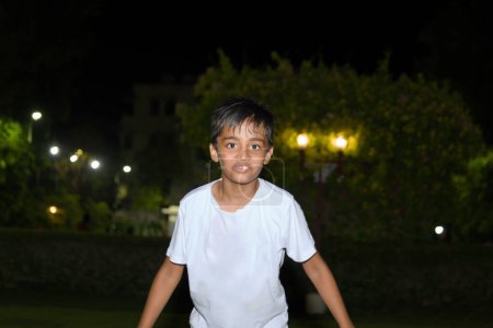 Un jeune garçon asiatique réfléchi regarde un parc illuminé par des lumières du soir.
