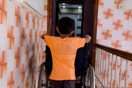 A young Asian boy with a determined expression stands beside his wheelchair at his front door