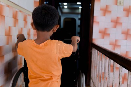 An image of a young Asian boy using a wheelchair, standing at the doorway of his home