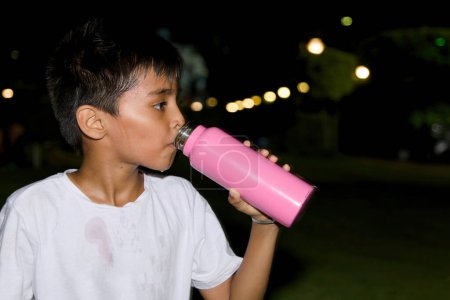 Ein junger asiatischer Junge nimmt ein erfrischendes Getränk Wasser aus seiner Flasche, während er sich nachts in einem Park entspannt.
