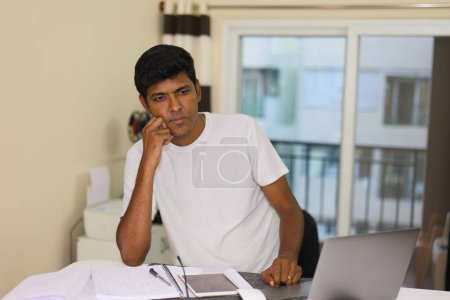 Ein indischer Mann wirkt nachdenklich und konzentriert, während er an seinem Laptop am Schreibtisch im Home Office arbeitet.