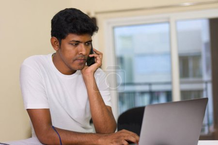 Un jeune Indien concentré jongle parfaitement avec les tâches, utilisant un ordinateur portable et parlant sur un téléphone dans son confortable bureau à la maison.