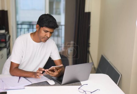 Concentrated young Indian man wearing headphones using laptop computer while working remotely at his home office.