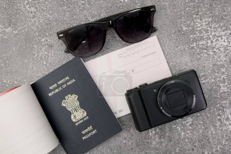 Cette image met en valeur les incontournables du voyage : passeport indien, appareil photo, lunettes de soleil et chèque, le tout disposé sur une surface plane en pierre grise.