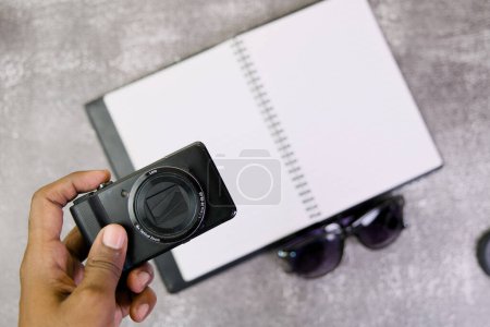 Eine Nahaufnahme über dem Kopf eines Fotografen mit einer Digitalkamera in der Hand, einem leeren Notizblock für Ideen und einer Sonnenbrille in der Nähe