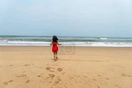 A young child in a bright red dress stands alone on a vast sandy beach, gazing out at the turbulent waves of the ocean