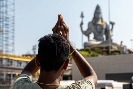 Une personne dans le traditionnel Anjali Mudra prie devant une statue majestueuse de divinité hindoue dans un temple serein. Les échafaudages de construction laissent entrevoir une rénovation en cours. murudeshwar