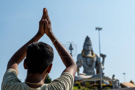 A person in traditional Anjali Mudra prays before a majestic Hindu deity statue at a serene temple. Construction scaffolding hints at ongoing renovation. murudeshwar