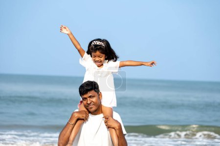A father carries his daughter on his shoulders, both smiling against the ocean backdrop. A cherished moment of love and play.