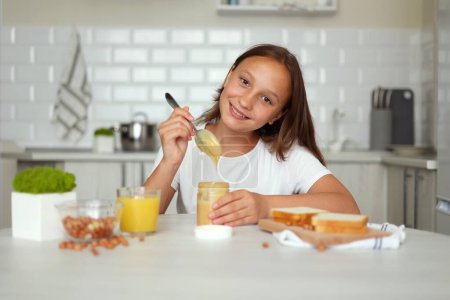 Téléchargez les photos : Une fille de bonne humeur faisant un sandwich au beurre d'arachide dans la cuisine. Photo de haute qualité - en image libre de droit
