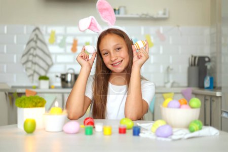 Téléchargez les photos : Une adolescente à l'intérieur d'une cuisine à la maison décorée pour Pâques décore les œufs de Pâques. Photo de haute qualité - en image libre de droit