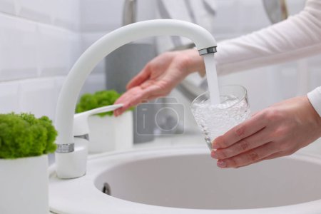 Téléchargez les photos : Une fille verse de l'eau propre dans un verre d'un robinet de cuisine. Eau purifiée. Photo de haute qualité - en image libre de droit