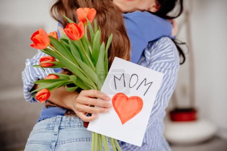 Photo for Grateful mom hugging daughter girl, holding flowers bouquet, receiving hand drawn greeting card with loving heart from girl, smiling at camera. Mothers day, 8 march, concept. Head shot portrait. Close-up of hands holding a bouquet of tulips and a han - Royalty Free Image
