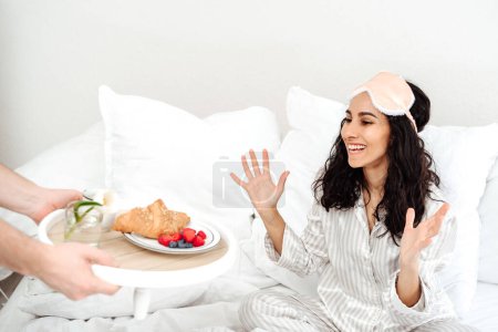 Photo for Beautiful woman of Eastern appearance rejoices in breakfast in bed. Hands holding a tray with croissant, berries and a flower. Girl in pajamas has just woken up and brightly rejoices at the surprise. - Royalty Free Image