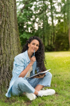 Photo for Portrait of attractive cheerful mixed race woman sitting on grass writing to-do list while spending free time outdoors. The girl sits in a lotus position in the park, thinks, holds a notebook - Royalty Free Image