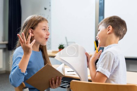 Photo for Lively conversation of children in the classroom. A schoolgirl explains to her desk mate how letters are written. The boy is interested and thoughtful. Creative communication of children as teacher. - Royalty Free Image