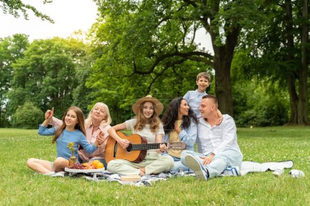 Foto de Picnic, gran familia descansando en la hierba para las vacaciones de verano, recreación al aire libre y estilo de vida saludable juntos. Abuelos, madre e hijos con una guitarra en un parque de campo - Imagen libre de derechos