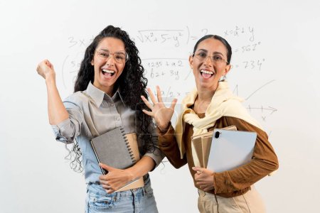 Photo for Two happy latin students with approved exams in the classroom. Joyful female students, shouting with delight, raised their hands up, holding school supplies in their hands. Girls stand at the board - Royalty Free Image