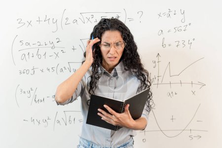 Freelance Stress. Concerned female Middle Eastern student, having problems with project, standing at board and looking at notebook, thinking about deadline and troubles. Curly girl holding her head