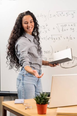 Young happy Eastern woman, professional teacher, online tutor standing at desk in front of chalkboard in classroom working on laptop computer teaching remote education virtual classes, vertical.