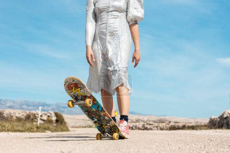 Foto de Una mujer con un atractivo vestido plateado mantiene su pie en un monopatín. En el fondo hay un hermoso cielo azul. - Imagen libre de derechos