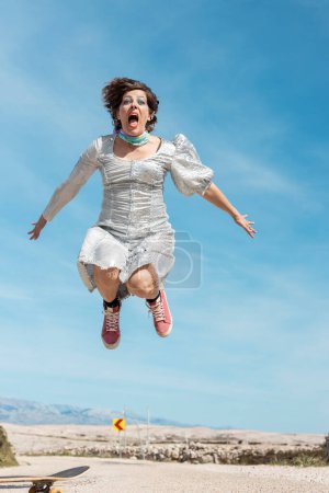 Foto de Una mujer feliz en un vestido de plata atractiva monta un monopatín y salta en el aire. En el fondo hay un hermoso cielo azul. Woman es la músico e intérprete croata Ane Paska. - Imagen libre de derechos