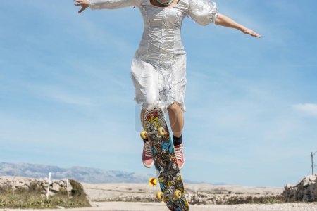 Foto de Una mujer con un atractivo vestido plateado monta un monopatín en la carretera abierta y salta al aire. En el fondo hay un hermoso cielo azul. - Imagen libre de derechos