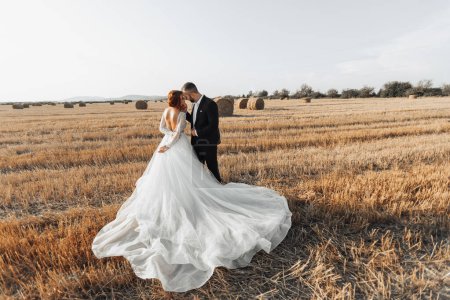 The bride and groom are standing in the field, and behind them are large sheaves of hay. The bride stands with her shoulders turned to the camera. Long elegant dress. Stylish groom. Summer